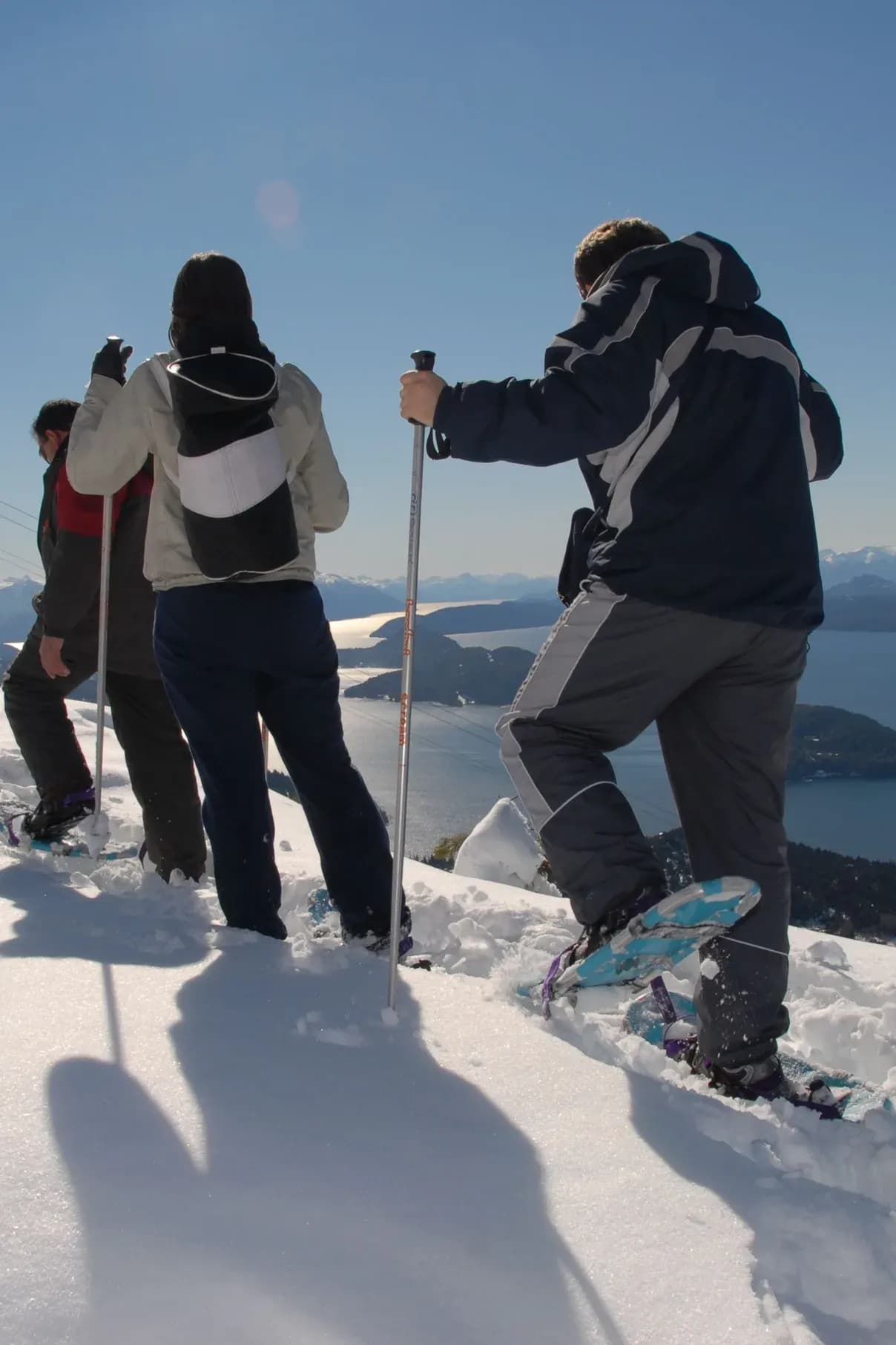 Three people with their backs to the camera walk with snowshoes and poles on a snowy hillside, taking in a panoramic view of a large blue lake dotted with islands and distant mountains under a clear sky.