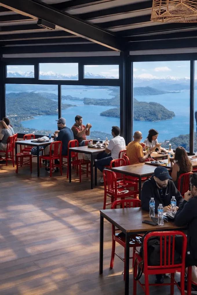 Praça de alimentação com pessoas comendo e admirando a vista panorâmica do Lago Nahuel Huapi.