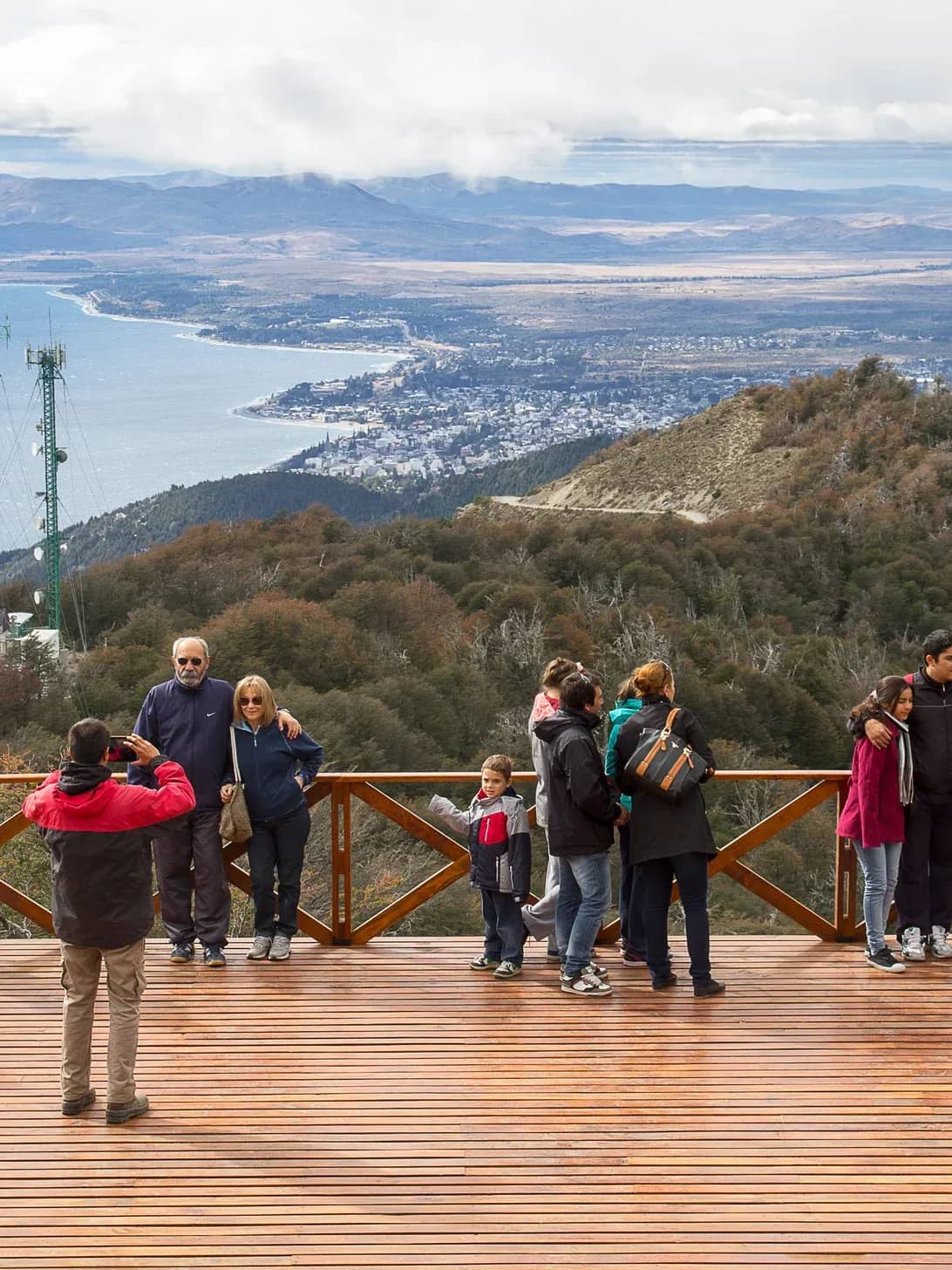 A cable car employee takes a photo of a group of people on a panoramic deck with Lake Nahuel Huapi and mountains in the background.
