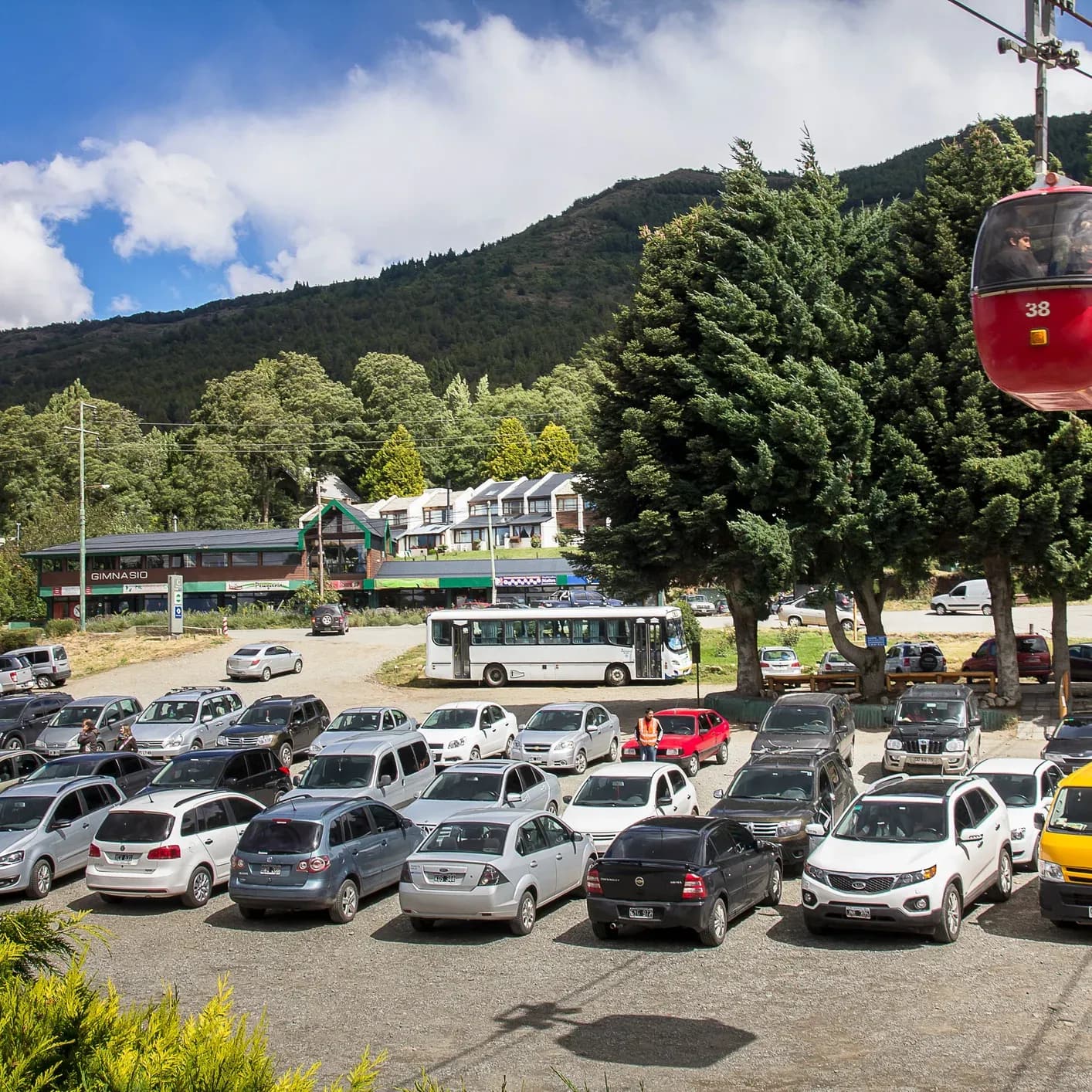Estacionamento na base do Teleférico Cerro Otto, com um teleférico subindo e carros abaixo.