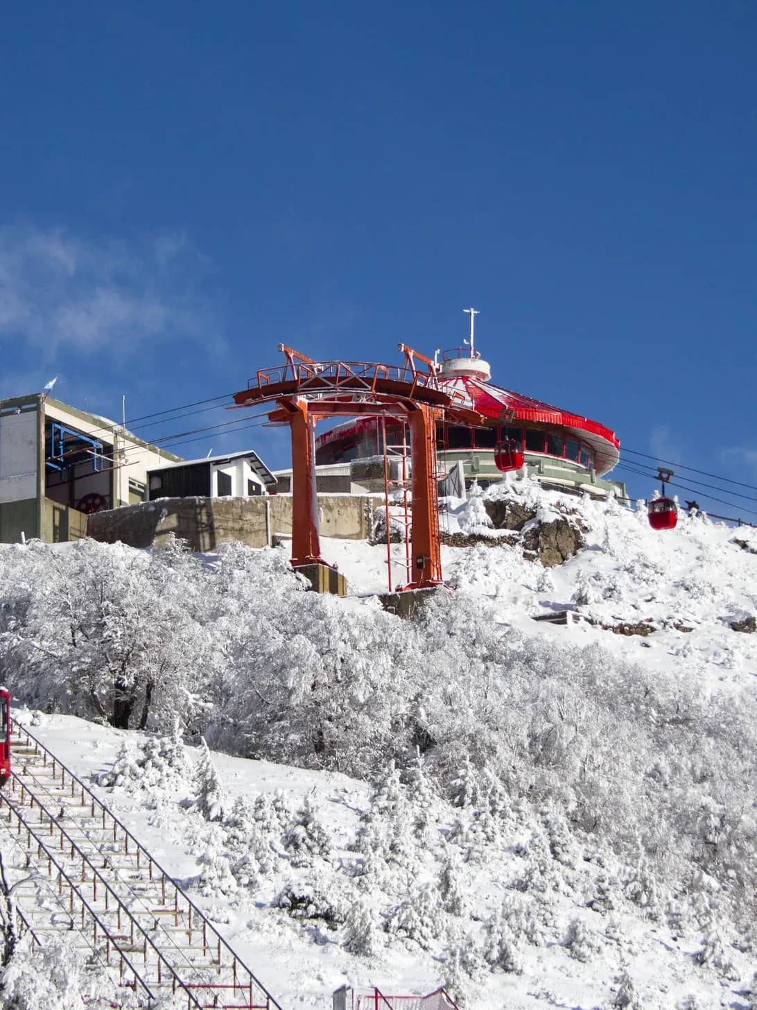 Vista panorâmica do topo do Cerro Otto, coberto de neve, mostrando a confeitaria giratória, a estação superior do teleférico e o funicular que leva ao topo.