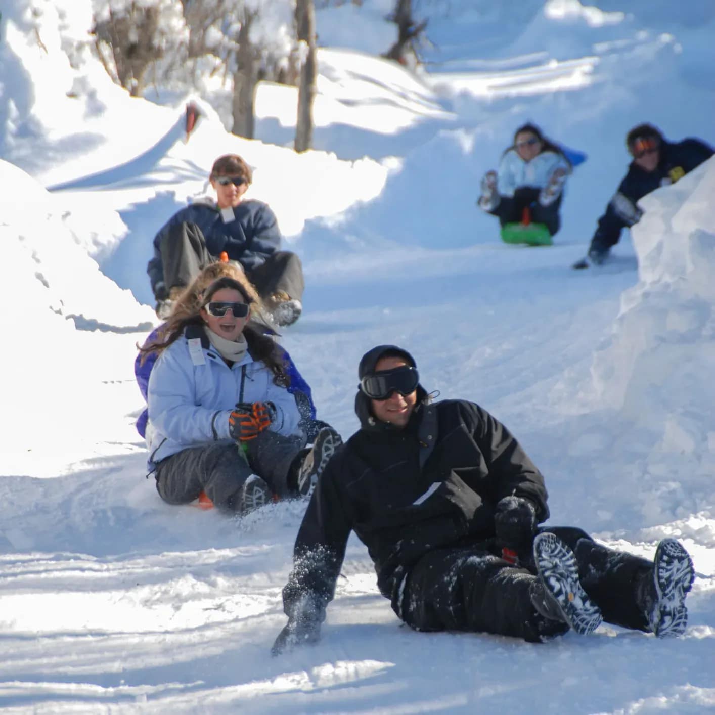 A group of friends going down a snow slope on individual sleds, laughing and expressing great excitement, with snow gear and helmets on a sunny winter day.