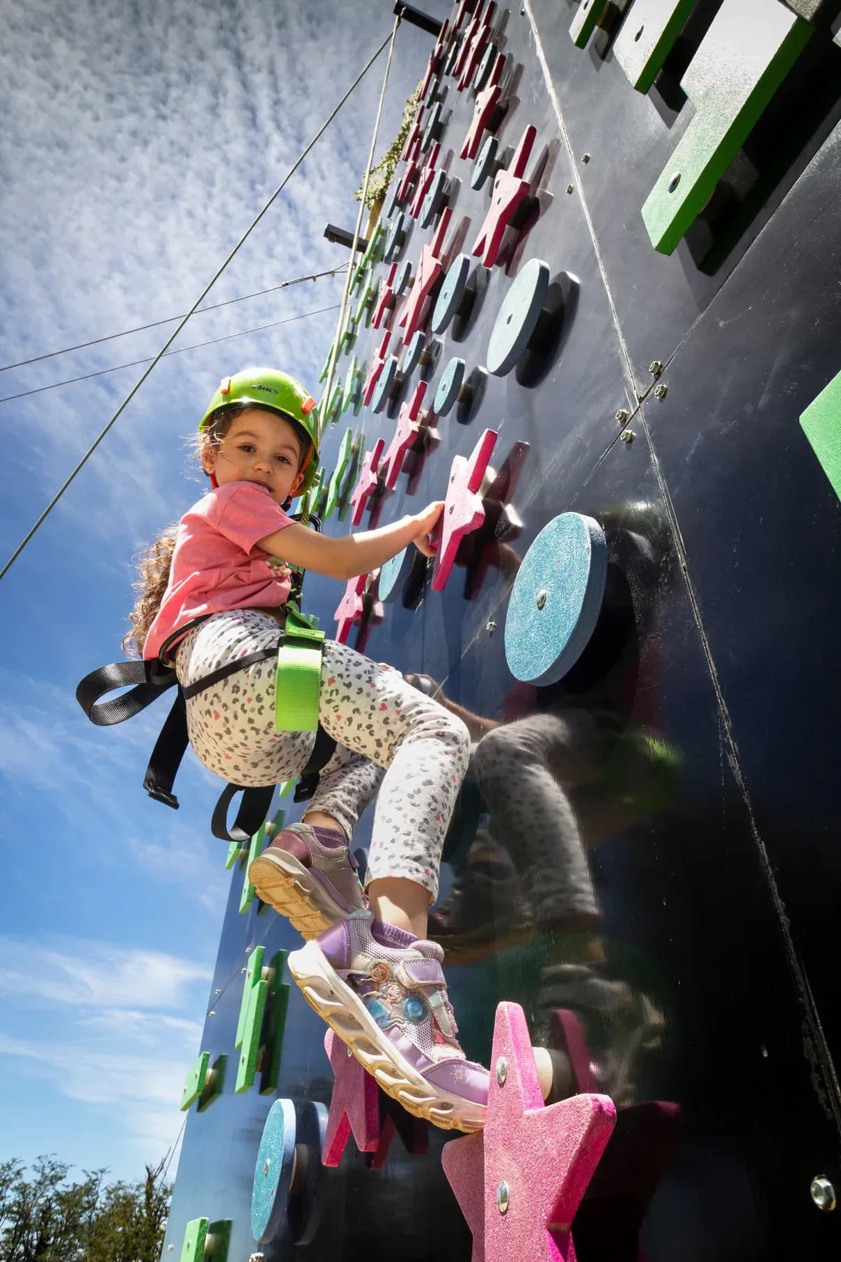 A young girl wearing a green hard hat and safety harness climbs a playful outdoor climbing wall. The wall has colorful holds shaped like stars and circles, and a blue sky with light clouds is visible in the background from a low angle.