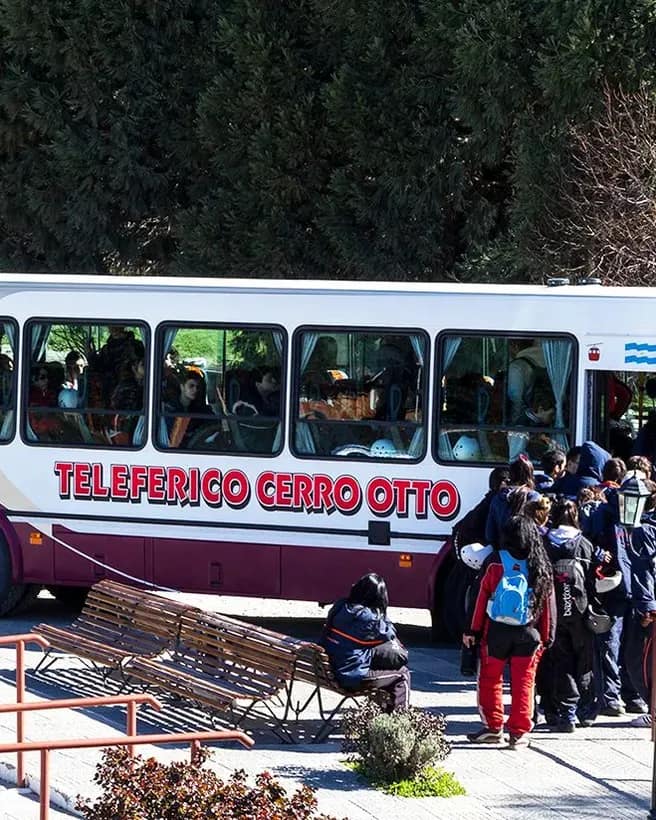 Ônibus turístico gratuito com o logotipo do teleférico Cerro Otto no estacionamento da base, com pessoas desembarcando.