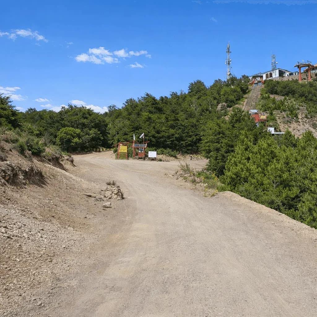 Gravel road at the summit of Cerro Otto overlooking the Teleférico complex, surrounded by forest and under a clear sky.