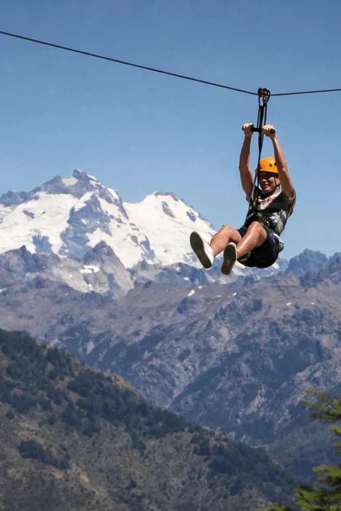 A man wearing an orange helmet glides down a zip line at great heights, with a stunning landscape of rugged mountains and snow-capped peaks under a clear blue sky.