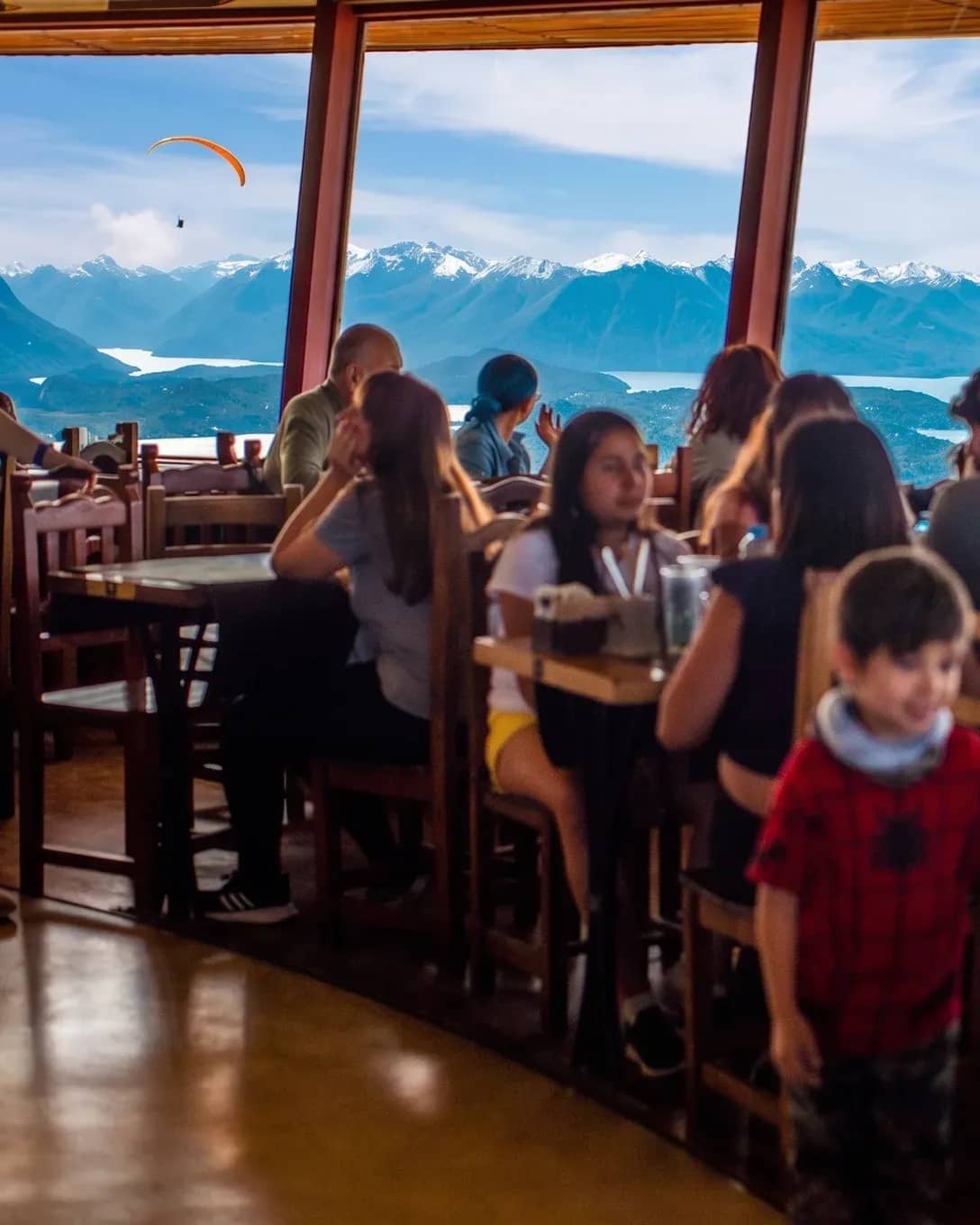 Pessoas almoçando dentro da Confeitaria Giratória com as montanhas e o Lago Nahuel Huapi ao fundo, visíveis através da janela.
