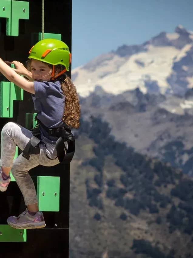 Girl wearing a green helmet and secured with a harness and rope climbing a climbing wall with a snowy mountain in the background